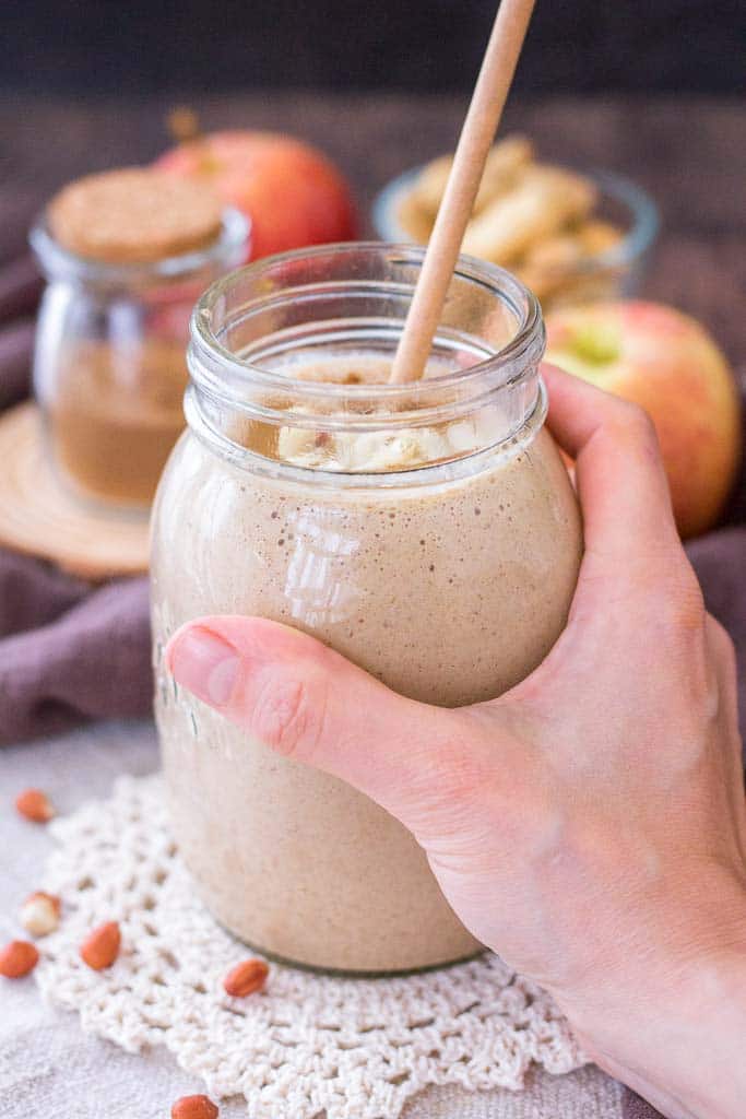 Hand holding a Apple Peanut Butter Smoothie served with a straw in a glass jar topped with creamy peanut butter and crushed peanuts