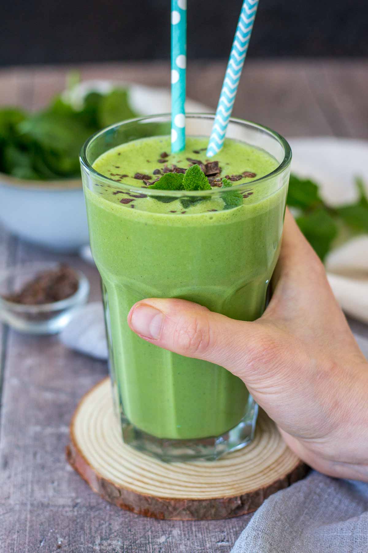 Hand holding shamrock protein shake served in a glass on a wooden coaster.